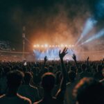 A vast night crowd with raised arms and phone lights facing a distant lit stage in an Indian stadium