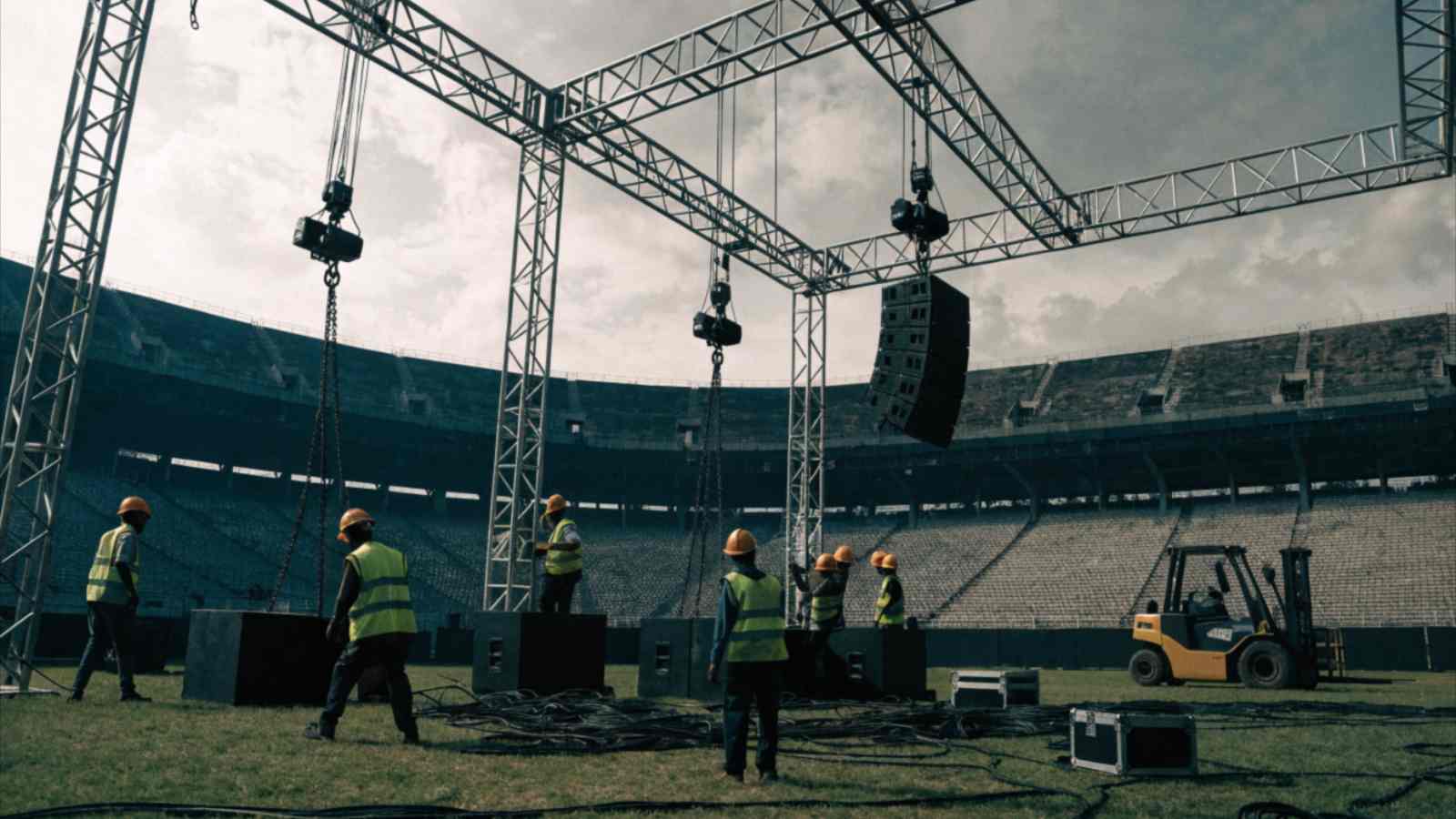 Crew in hard hats rig steel truss and speaker arrays on a stage built inside an empty stadium