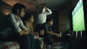 An Indian family crowded around a large television watching a floodlit cricket match at night
