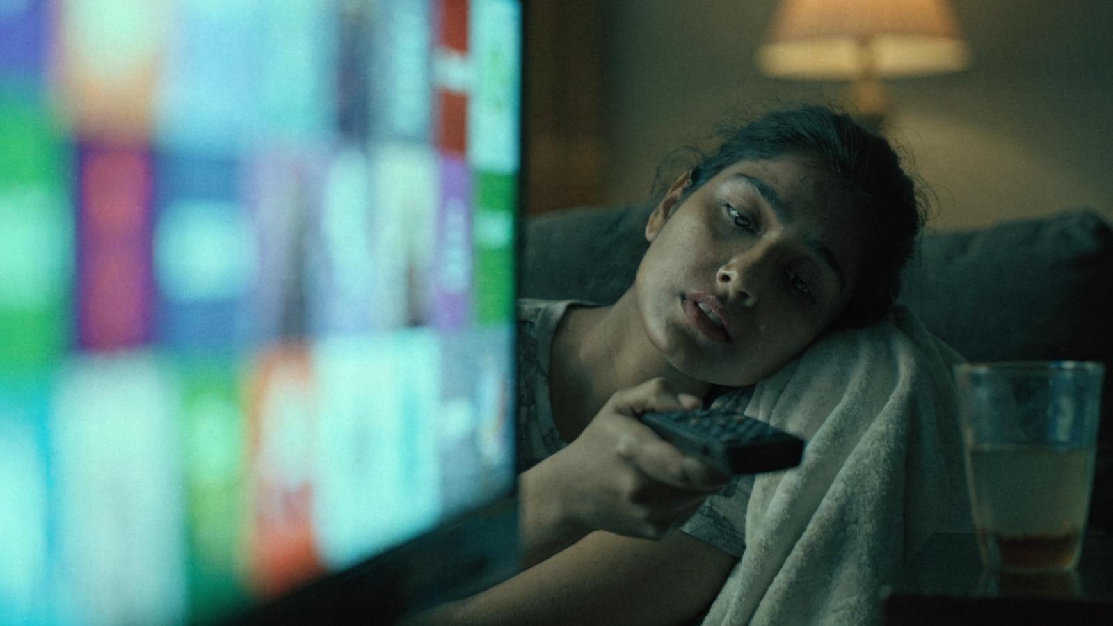 A young woman alone on a sofa at night holding a remote, looking bored at a grid of blurred tiles on the TV