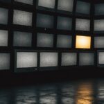 A wall of televisions showing static, one screen still glowing
