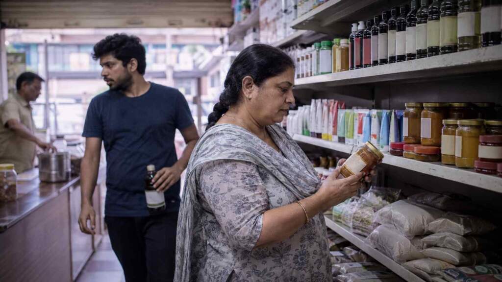 A woman reads the label on a jar in an Indian general store while a younger shopper walks away holding a bottle