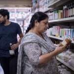 A woman reads the label on a jar in an Indian general store while a younger shopper walks away holding a bottle