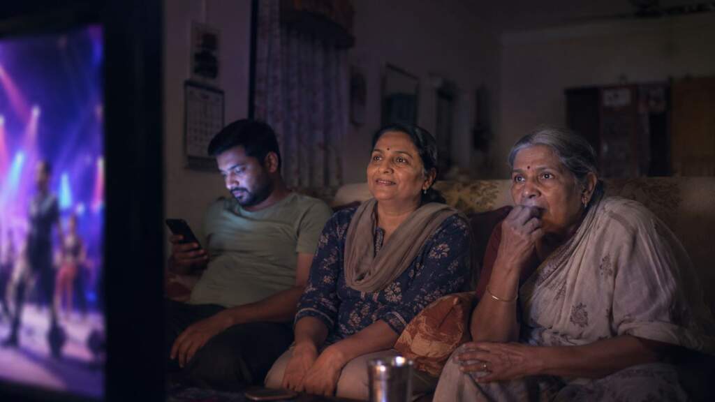 An Indian family watches a reality show at night: an older woman absorbed, a woman smiling, a younger man on his phone