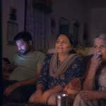 An Indian family watches a reality show at night: an older woman absorbed, a woman smiling, a younger man on his phone