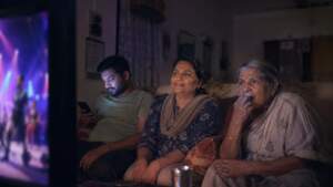 An Indian family watches a reality show at night: an older woman absorbed, a woman smiling, a younger man on his phone