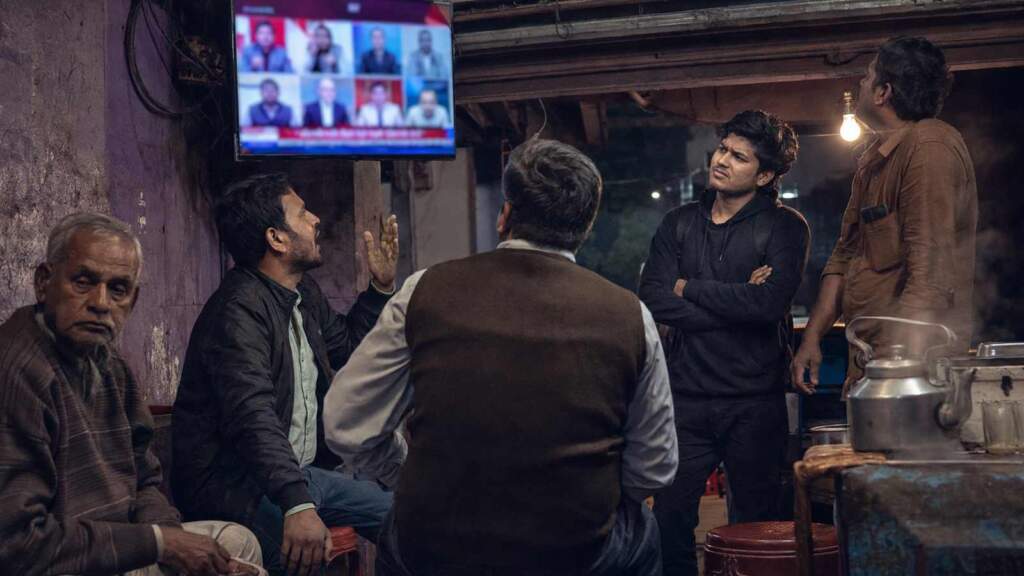 Men of different ages watch a Hindi news debate at a tea stall at night, reacting differently to the screen