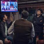 Men of different ages watch a Hindi news debate at a tea stall at night, reacting differently to the screen