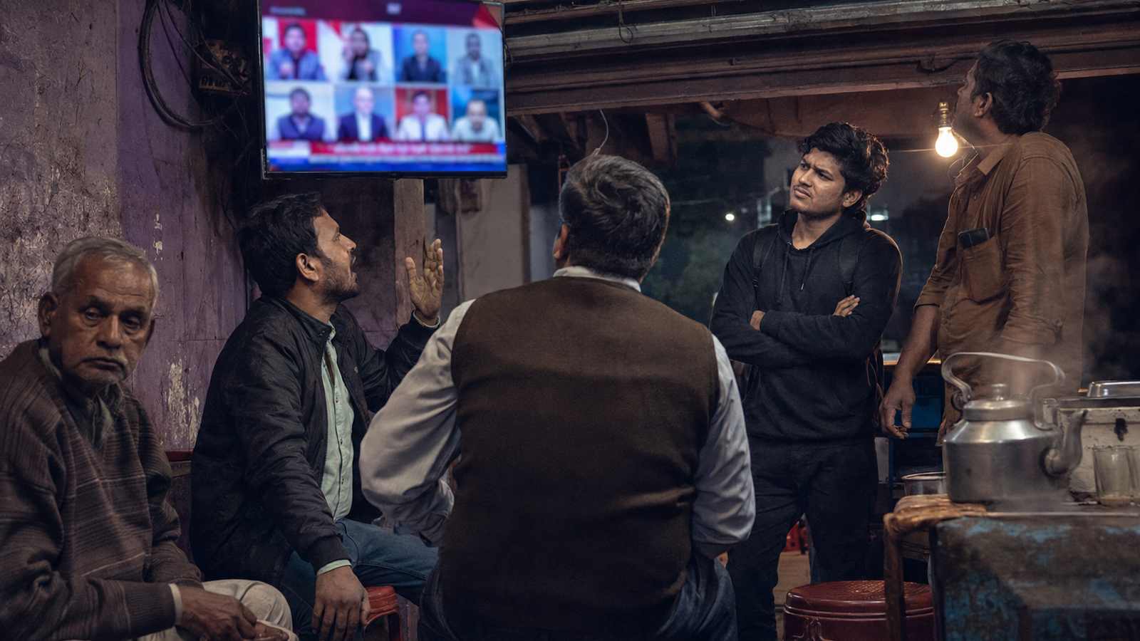 Men of different ages watch a Hindi news debate at a tea stall at night, reacting differently to the screen