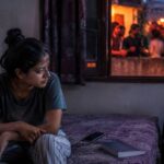 A young woman sits alone on her bed at dusk while friends talk on a lit terrace outside her window