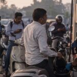 A rider on a scooter watches the fuel nozzle as an attendant fills his tank at a crowded petrol pump at sunrise