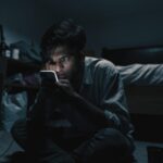 A young man on the floor of a dark room lit only by his phone screen