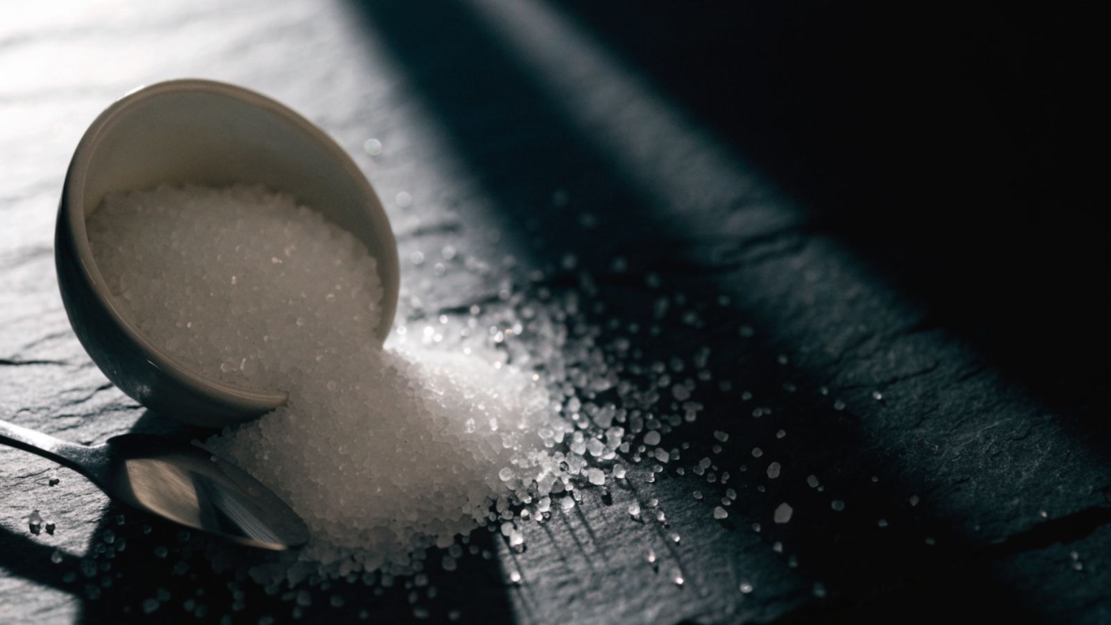A bowl of table salt spilling across dark slate under raking light