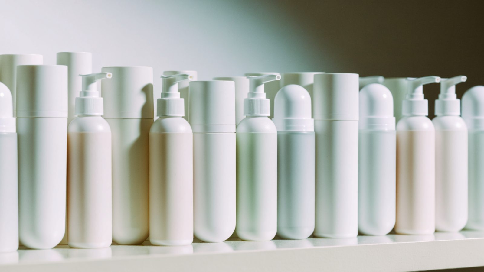Plain white and pastel cosmetic tubes and bottles on a pale shelf